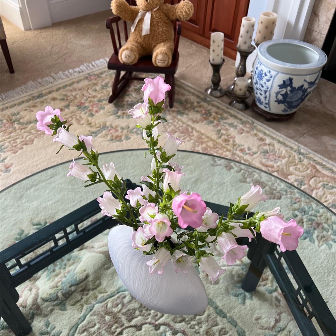Vase with pink Canterbury Bells on a glass table, background includes a teddy bear and decorative items.