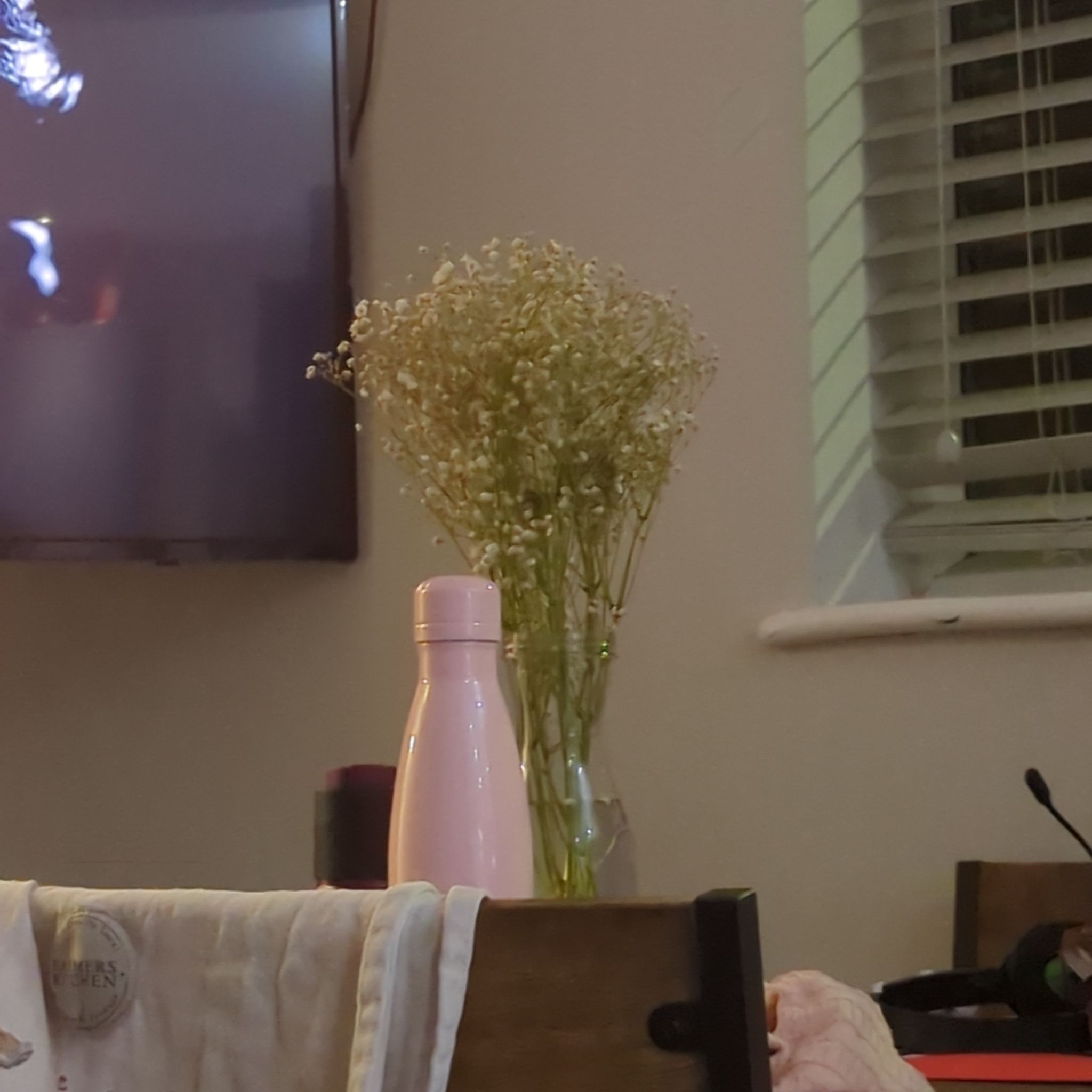 Vase with Baby's Breath flowers on a table, background includes household items and a window.